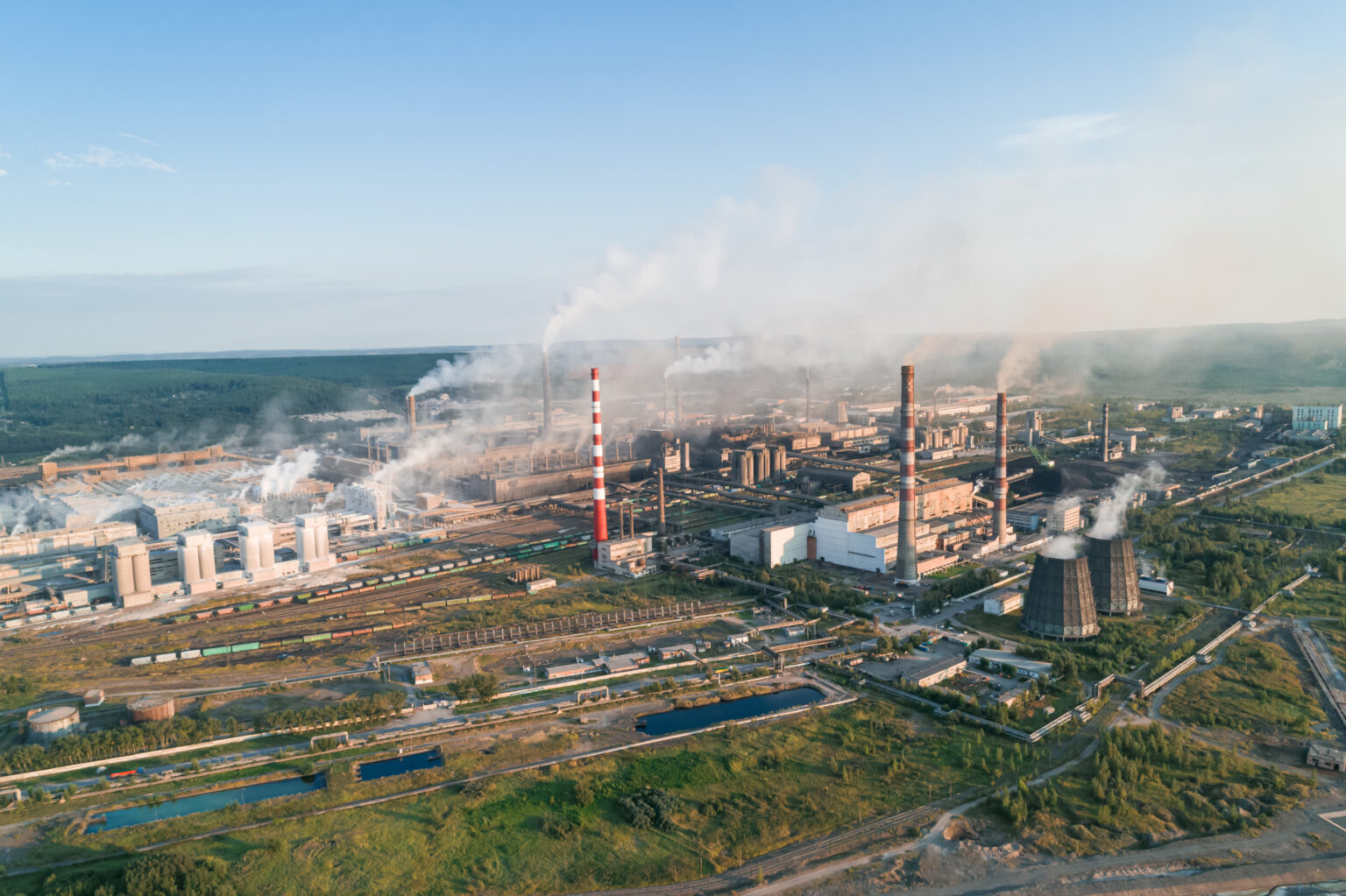 Toxic smoke rises from multiple industrial plants, filling the air above the landscape with pollutants as the sun sets in the background, highlighting serious environmental issues.
